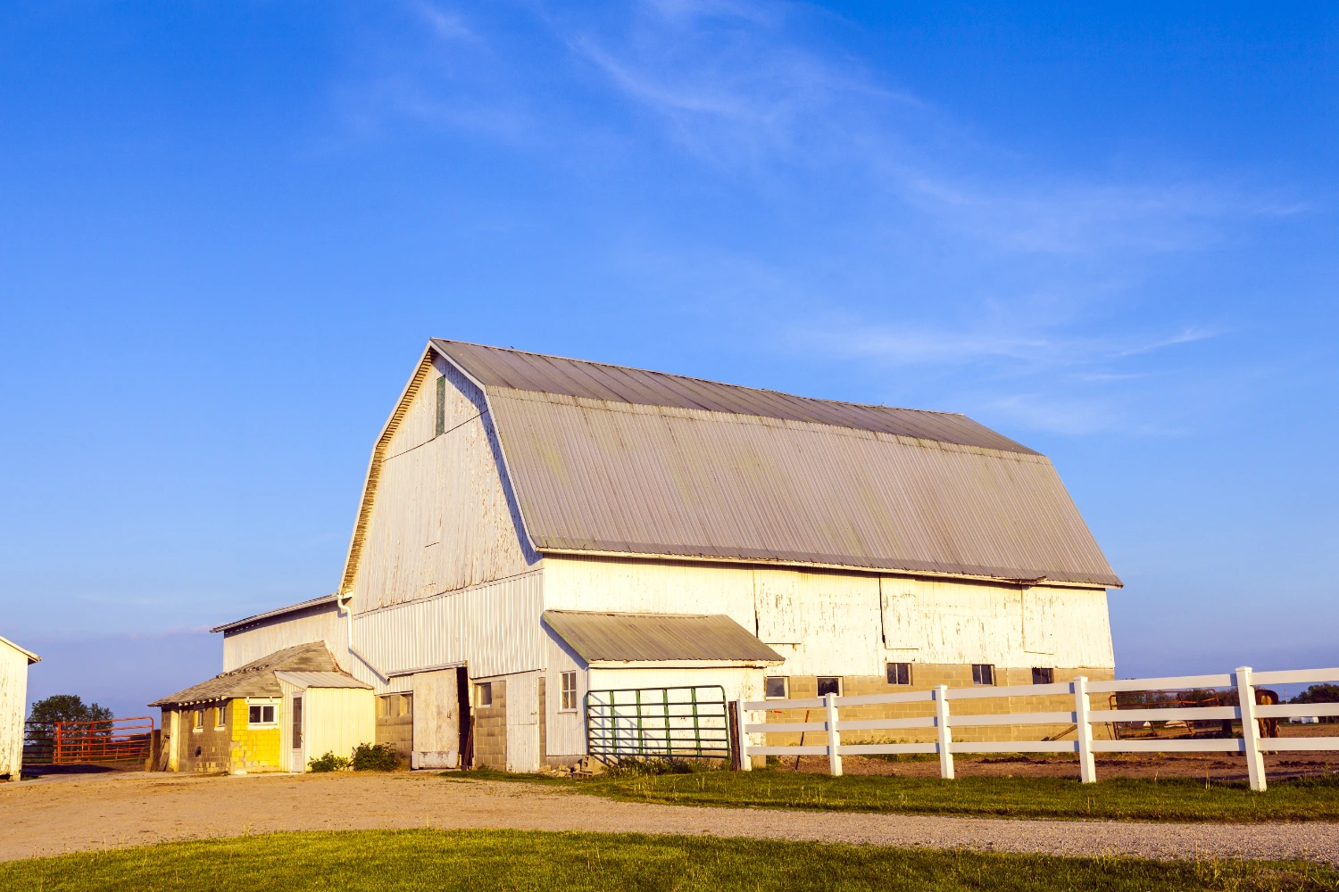 White gambrel-roof barn with a weathered metal roof and a yellow block annex, glowing in golden evening light behind a white fence under a bright blue sky. in Midland, TX